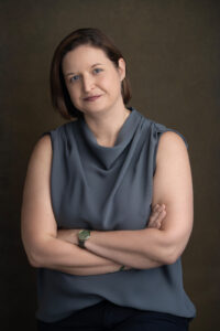 A woman with short brown hair crosses her arms. She is wearing a gray sleeveless shirt.